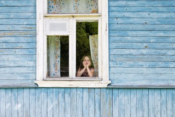 Sad bored little girl looking out the country house window leaning her face on her hands. Outside view