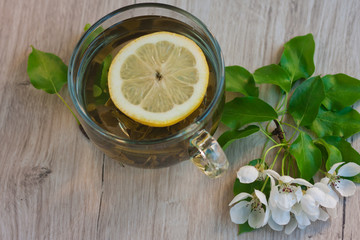 A large cup of fruit tea with lemon, a glass cup with jam and a flowering branch near. A blossoming branch of an apple tree. A small jar of jam with a spoon
