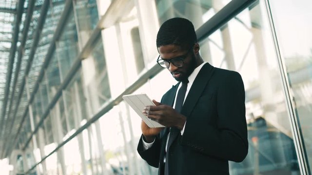 Happy african american businessman using tablet device while standing near big office windows outdoors. Man scrolling pages, tapping on touchscreen, checking financial charts. close up