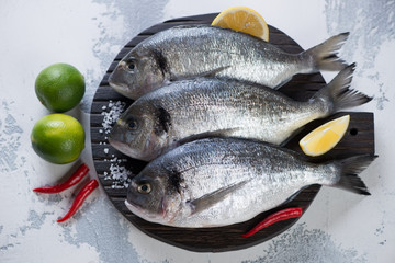 Wooden serving board with fresh uncooked dorado fish and seasonings. High angle view on a white concrete background