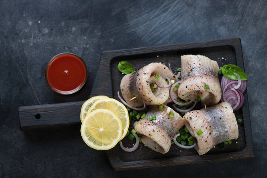 Black wooden serving tray with herring fillet rolls, flat-lay over scratched metal background, horizontal shot