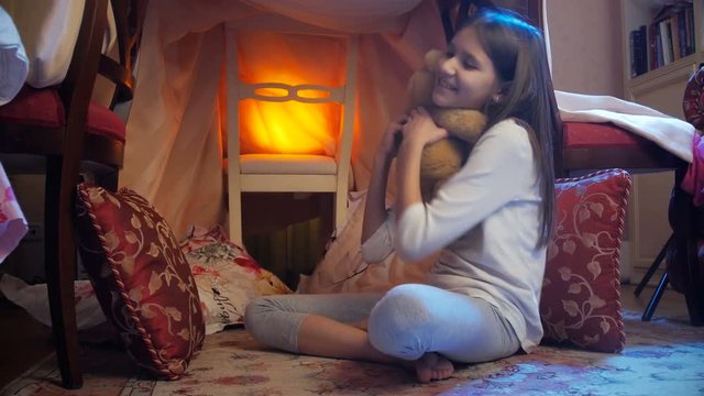 Dolly shot of cute smiling girl in pajamas playing with teddy bear in house made of blankets