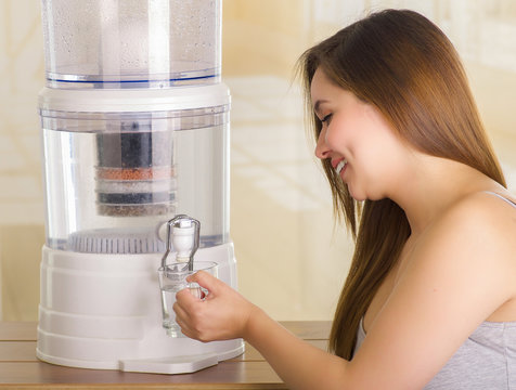 Close Up Of A Beautiful Smiling Woman Filling A Glass Of Water, With A Filter System Of Water Purifier On A Kitchen Background