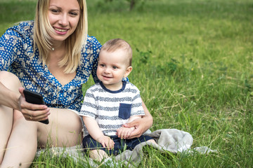 Young pretty mather and a baby boy sitting on grass and looking at electronic device