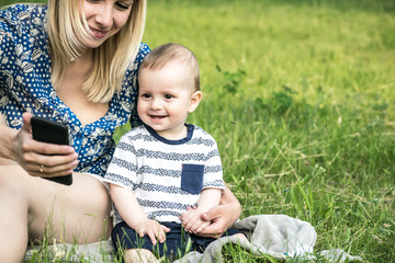 Young mather and a baby boy sitting on grass and looking at a Smartphone