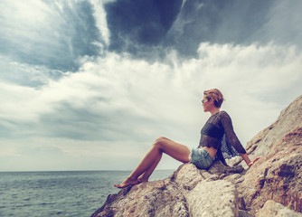 Young beautiful stylish woman sitting on a stone by the sea and looking at the horizon