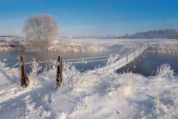 Real Russian Winter.Winter Landscape With Trail Across The Rural Suspension Bridge Over The Foggy River,Reed Covered With Hoarfrost And Traditional Wooden Houses On Snow-Covered Field In Sunny Day- 158332317