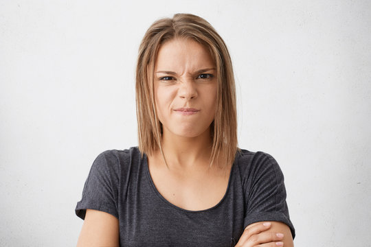 Angry young female with dark eyes frowning her nose and lips wearing casual T-shirt standing crossed hands isolated over white background showing her dissatisfaction. People and emotions concept