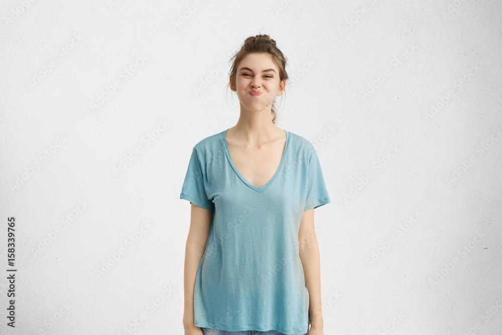 Wall mural Portrait of playful funny teenage girl wearing blue t-shirt having fun indoors, holding breath, doing her best not to burst into laughter while friends trying to make her laugh. Human emotions