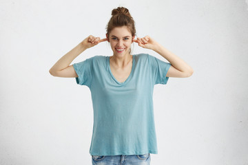 Beautiful teenage girl wearing jeans and oversize blue t-shirt plugging ears with fingers and smiling broadly, teasing, pretending not to lsiten, ignoring someone. Human emotions and feelings