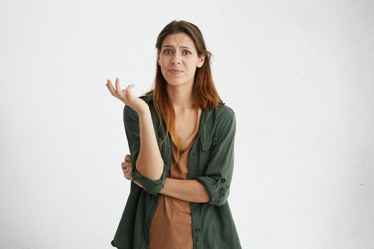 What? Casually dressed puzzled female standing at blank studio wall, having questionable look, her facial emotions and gesture expressing indignation, dislike and uncertainty towards something