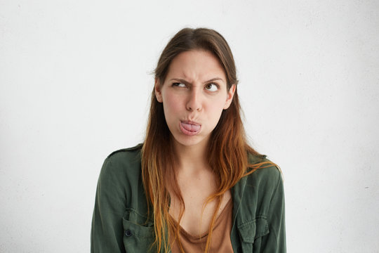 Crazy attractive woman showing her tongue frowning eyes looking aside having joy. Caucasian female standing against white wall blowing her cheeks showing tongue being angry showing her discontent