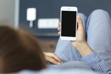 Back view of young female holding modern smartphone lying at comfortable bed in cozy hotel communicating with friends online. Close-up female hands holding modern device with copy screen space