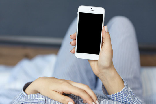Front view of female hands holding smartphone while lying in bed having rest watching videos online with copy space for your promotional content. Woman doing online shopping at home in bedroom