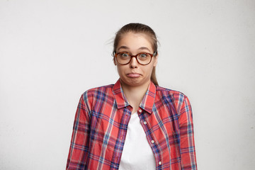 Portrait of puzzled young Caucasian pretty woman in casual clothes and eyeglasses shrugging her shoulders and screwing up her lips having surprised look. Cute female having no idea what to do