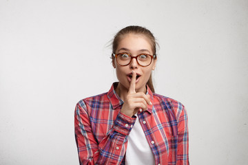 Don't tell anyone! Attractive young European female wearing hair in ponytail, keeping index finger at her lips, hushing, asking to hold tongue and not unlock her secrets, having excited look