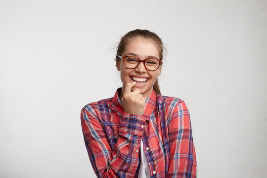 Portrait of attractive pleasant smiling woman with fair hair wearing elegant eyeglasses and checked red shirt holding finger on teeth isolated over white background having good mood after party.