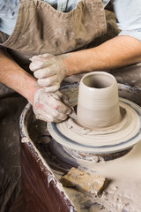 Fototapeta na wymiar pottery, workshop, ceramics art concept - hands of a man works with potter's wheel, fingers form shape of raw fireclay, male master sculpt a utensil with a sponge and carving tool, top view, vertical
