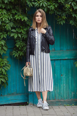 Young beautiful girl in stylish streetwear black leather jacket long striped dress white sneakers and with a fashionable bag posing against turquoise wooden doors