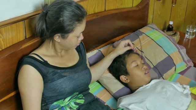 Medium high angle view of Asian mother taking care of her sick son . She strokes his hair, then takes his temperature with a digital thermometer and reacts. One of a series 