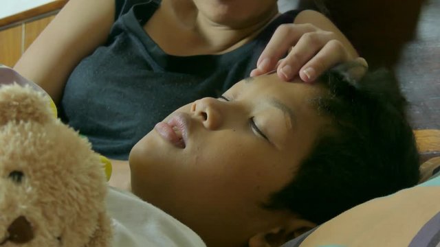 Medium close up of sick multicultural boy lying in bed as his Asian mother takes care of him, stroking his forehead. One of a series of multicultural lifestyle and people clips 

