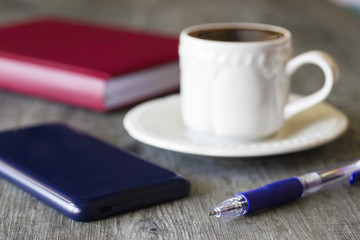 Objects on gray background. A white cup of coffee, pen, red notebook and blue cell.