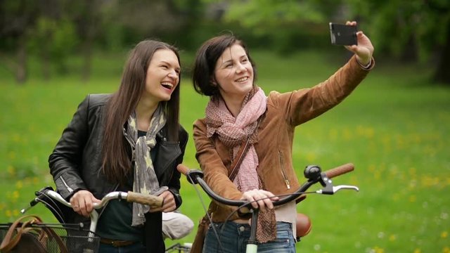 Two Female Bicyclists in Jackets and Scarves are Taking Photo by Smartphone. Young Beautiful Brunettes with Bicycles are Photographing Themselves Using Mobile Phone.