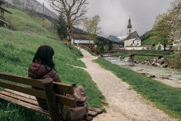 Germany. Ramsau. The girl traveler rests on a bench near the  Ramsauer Ache river overlooking the Parish Church of St. Sebastian