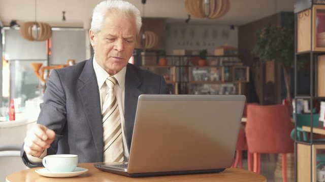Senior gray businessman looking at the laptop screen at the cafe. Aged caucasian man dressed in suit and tie making a pause to drink coffee. Attractive male manager analizing his work on computer