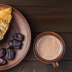 Hot chocolate or cocoa drink in cup and sweet croissant and dates fruit at plate on dark brown wooden table, top view
