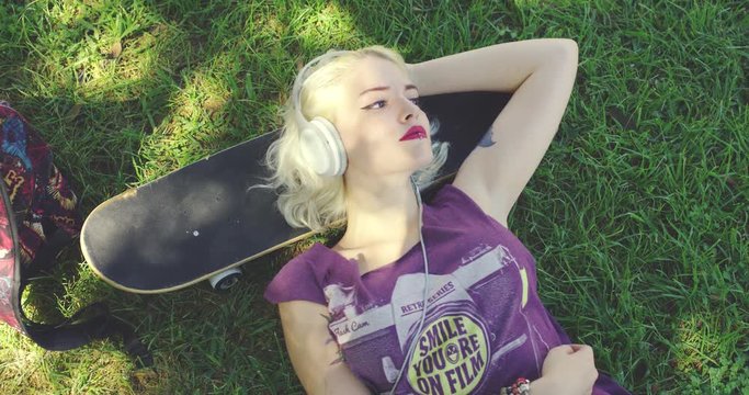 Pretty young blond woman relaxing on lush green grass in the shade of a tree in summer listening to music lying with her head on her skate board staring dreamily up into the air.