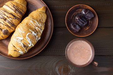 Hot chocolate or cocoa drink in cup and sweet croissants and dates fruit at plate on dark brown wooden table, top view