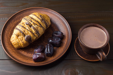 Hot chocolate or cocoa drink in cup and sweet croissant and dates fruit at plate on dark brown wooden table