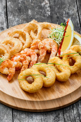 Seafood platter with deep fried squid rings, shrimp and onion rings decorated with lemon on cutting board on wooden background. Mediterranean appetizers. Top view
