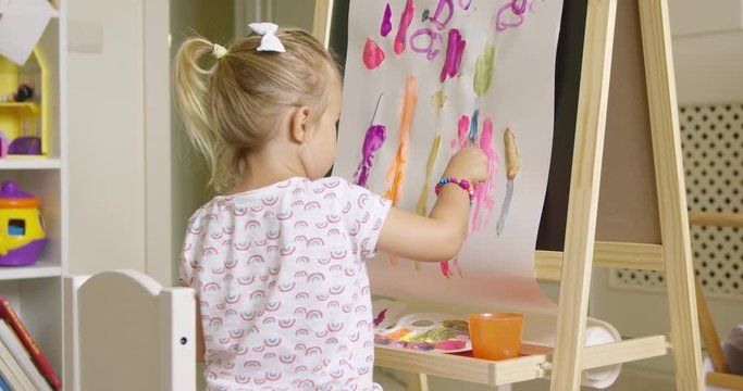 Cute creative little girl artist painting at an easel sitting applying brightly colored paints in an abstract design
