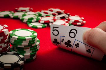 Hand holding five and six suited in clubs at a poker table surrounded by casino chips. 5 6 of suit is a bonus hand in many casino