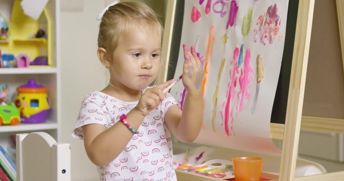 Cute little girl artist creating a colorful abstract painting on a sheet of paper attached to a blackboard in her playroom at home dabbing paint on her hand for a palm print.