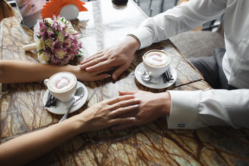 Fototapeta premium Wedding couple at cafe. Man holds woman's hand, drinks cappuccino. Bride and groom coffee break dating gift, bouquet on table