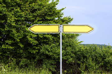 Empty yellow road signs pointing in opposite directions against green bushes and a blue sky, symbol...