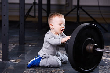 Little cute boy in sportwear sitting on the floor at the gym and looking on the barbell.