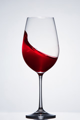 Splashing red wine with wave in the elegant wineglass standing against light background with reflection.