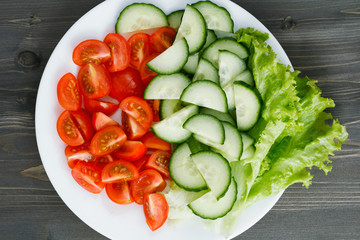 Salad of fresh cherry tomatoes, cucumber and lettuce leaves on a white plate on a dark wooden table. View from above.