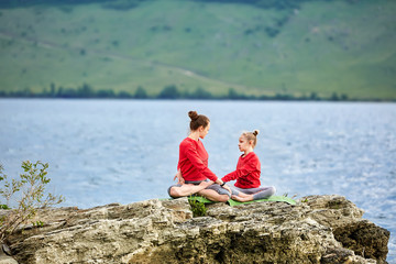 Young mother and daughter doing yoga exercises on the rock near river.