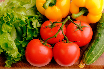 Branch of fresh red tomatoes, yellow bell pepper, green cucumbers and lettuce on a wooden cutting board