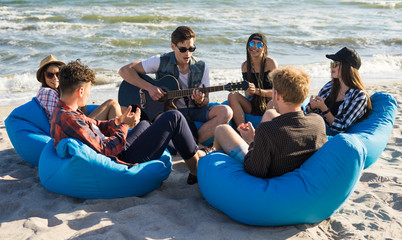 group of friends with guitar on the beach party - Young hipster people on summer vacation sitting on bean bags and playing guitar near the sea