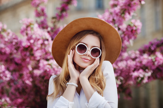 Outdoor close up portrait of young beautiful happy girl posing in street, near blooming tree. Model wearing stylish round sunglasses, yellow hat, wrist watch, white shirt. Female fashion concept