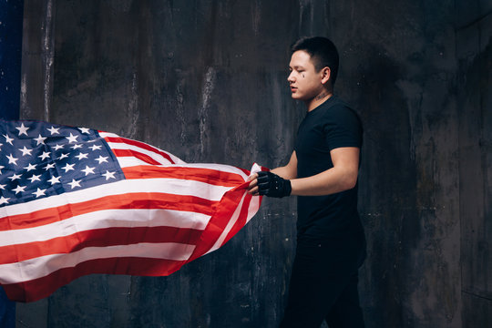 Young USA patriot with flying national flag. Strong man in black cloth with tattoo on dark studio background. Independence day, confidence, pride, fidelity to the nation, memorial day concept