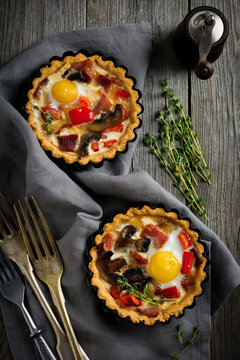 Mini tart with baked ham, pepper, mushrooms and quail eggs in creamy sauce on a wooden stand and old background. Selective focus.Top view.