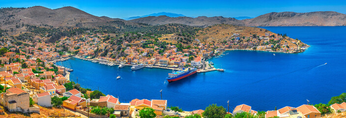 Panoramic view on beautiful Symi island Aegean Sea blue bay Greek houses on green hills, yacht sea port, tourist ferryboat. Famous Mediterranean MSC cruises. Greece islands holiday vacation tours trip - obrazy, fototapety, plakaty