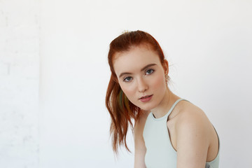 Portrait of gorgeous European teenage girl with green eyes and perfect clean skin wearing red hair in ponytail, standing isolated against white studio wall background, looking seriously